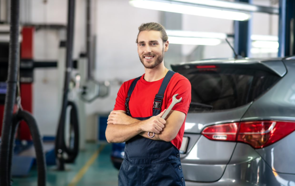 An auto repair shop technician wearing a red shirt is smiling and holding a wrench.